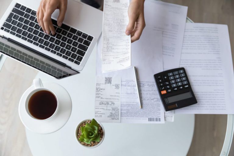 Mesa vista de cima com close nas mãos de uma pessoa acessando um notebook e segurando um documento. Na mesa há uma calculadora, uma xícara de café e várips outros documentos.