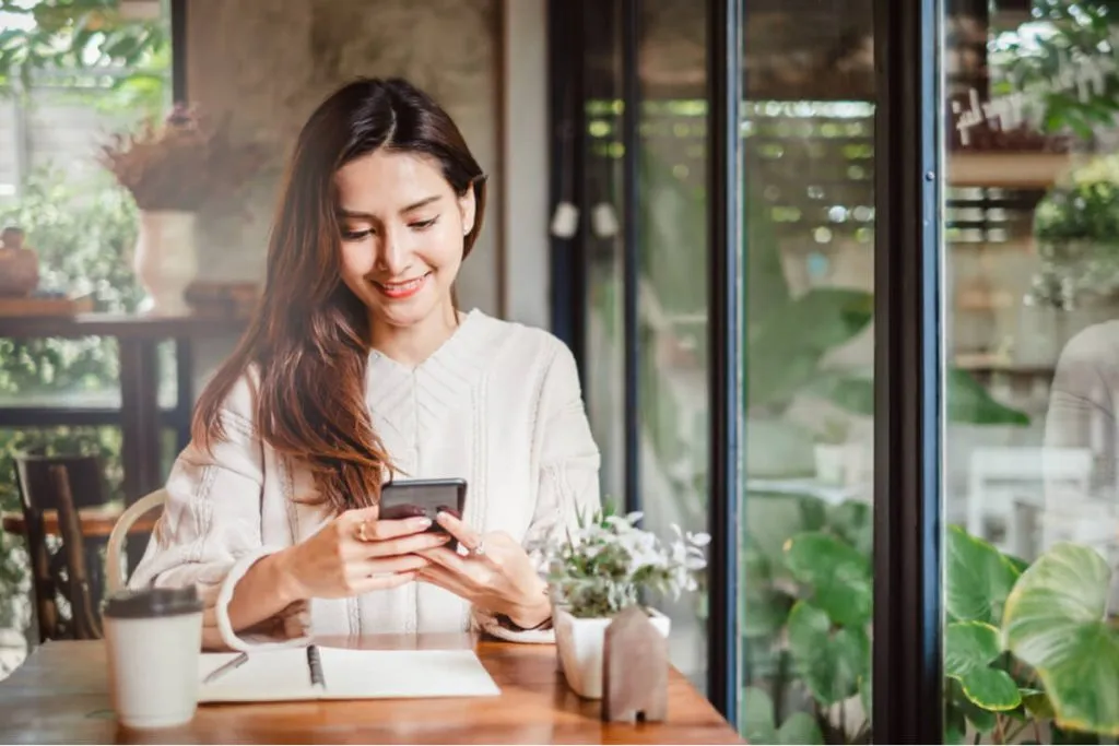 Uma jovem com semblante sorridente segurando um smatrphone e sentada à mesa em um dia claro, de forma descontraida.