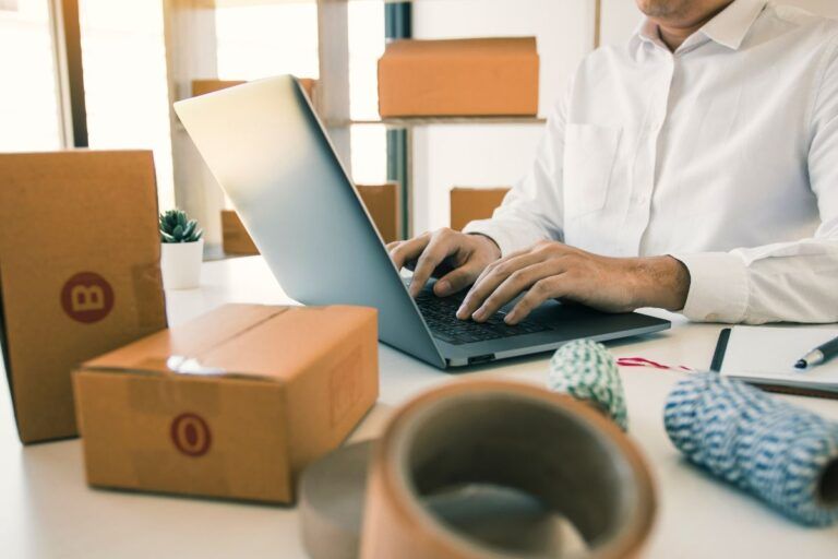 Homem acessando o Amazon Seller Central no notebook com caixas ao seu lado.