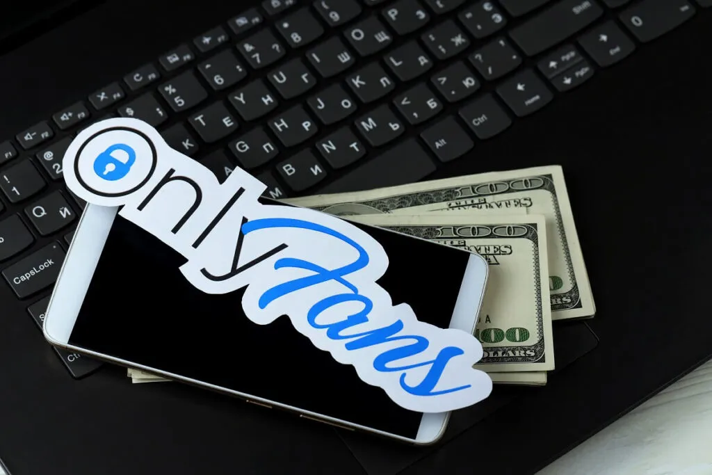 Logo do OnlyFans em cima de um celular, notas de dólar e por último um teclado de um notebook