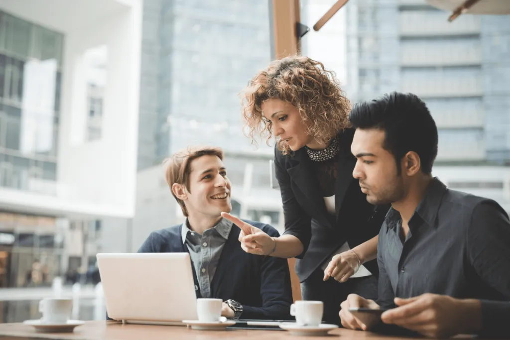 Dois homens sentados em frente a um notebook com uma mulher em pé junto a eles conversando