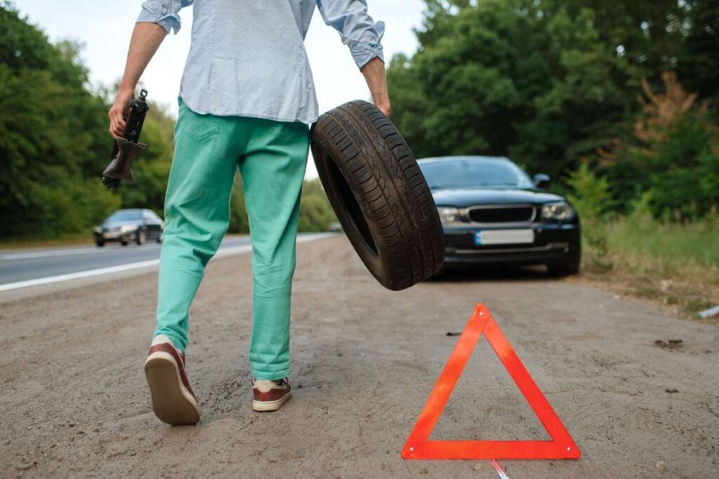 Homem segurando o pneu com o carro quebrado no fundo. 