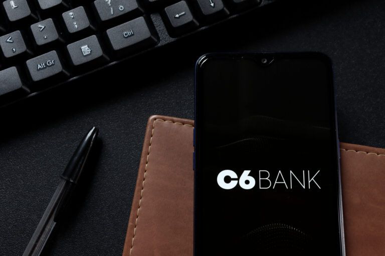 celular com logotipo do C6 bank na tela apoiado em cima de uma cardeneta com uma caneta e um teclado de computador ao lado