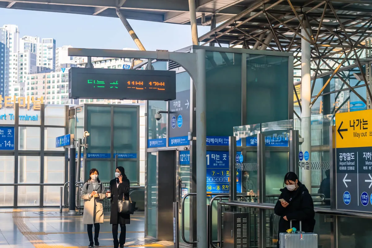 Três mulheres vestindo roupas de frio estão em uma estação de transporte. Duas caminham, uma fala ao celular, a outra segura um copo de café, enquanto a terceira está parada mais a frente mexendo no celular