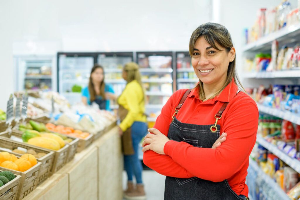 Atendente de mercado com uniforme, sorrindo em frente a seção de hortifruti, em comemoração ao Dia do comerciário.