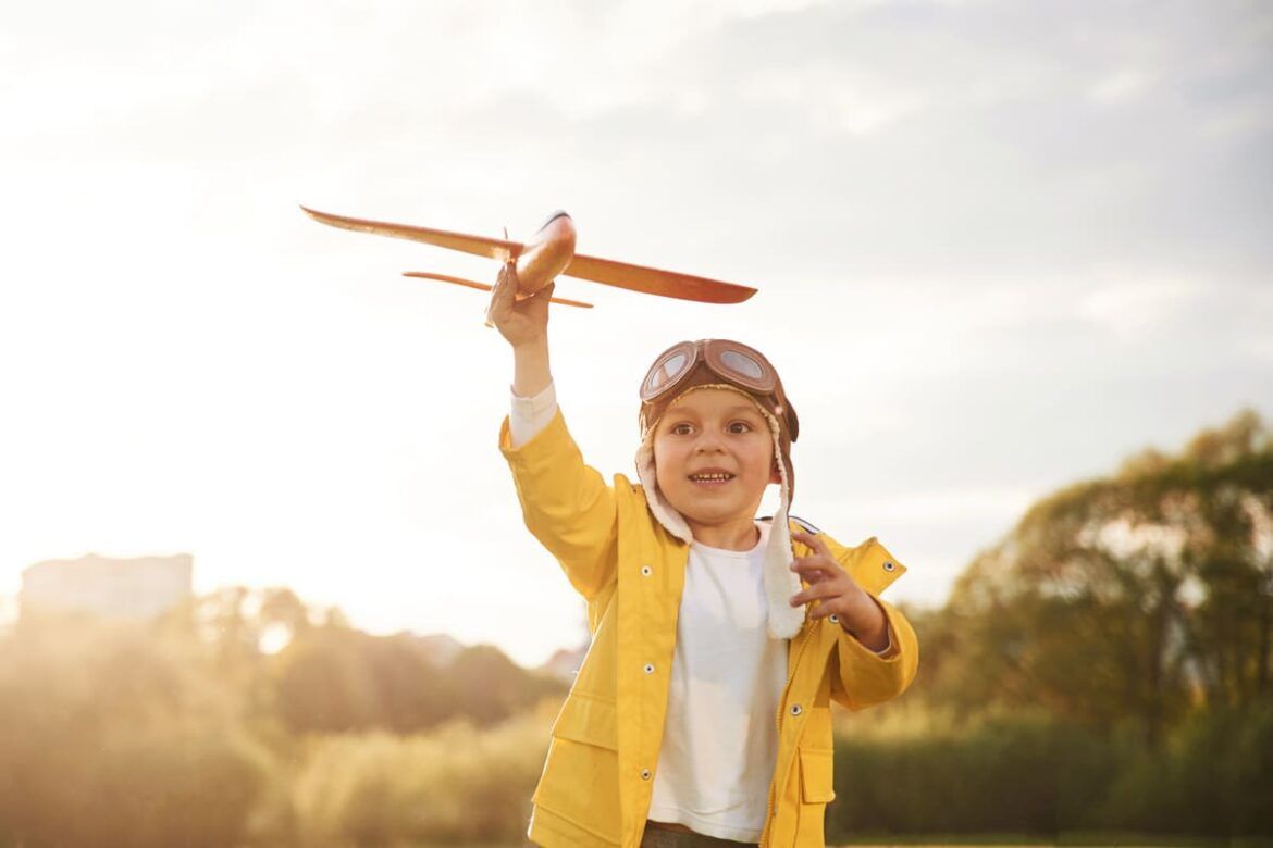 Criança sorridente brincando ao ar livre com um avião de brinquedo, vestindo um casaco amarelo e óculos de aviador retrô.