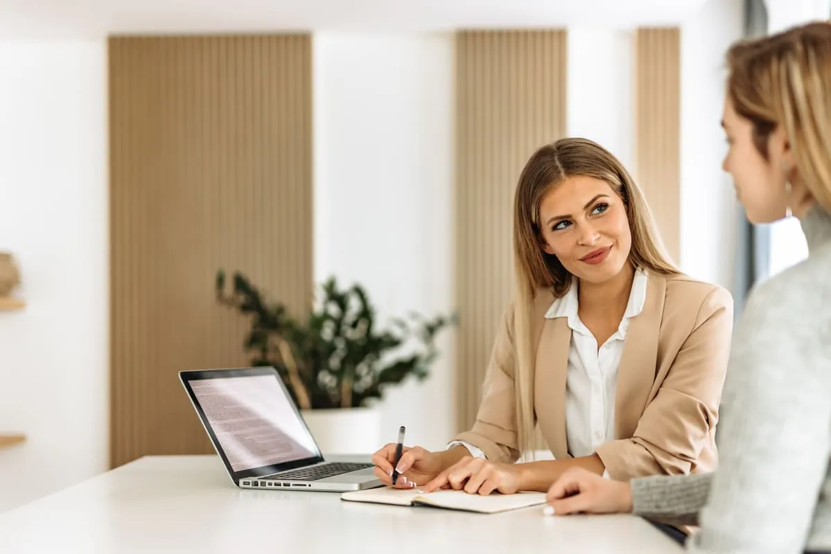 Chefe conversando com funcionária em uma mesa de escritório com um laptop ao lado.