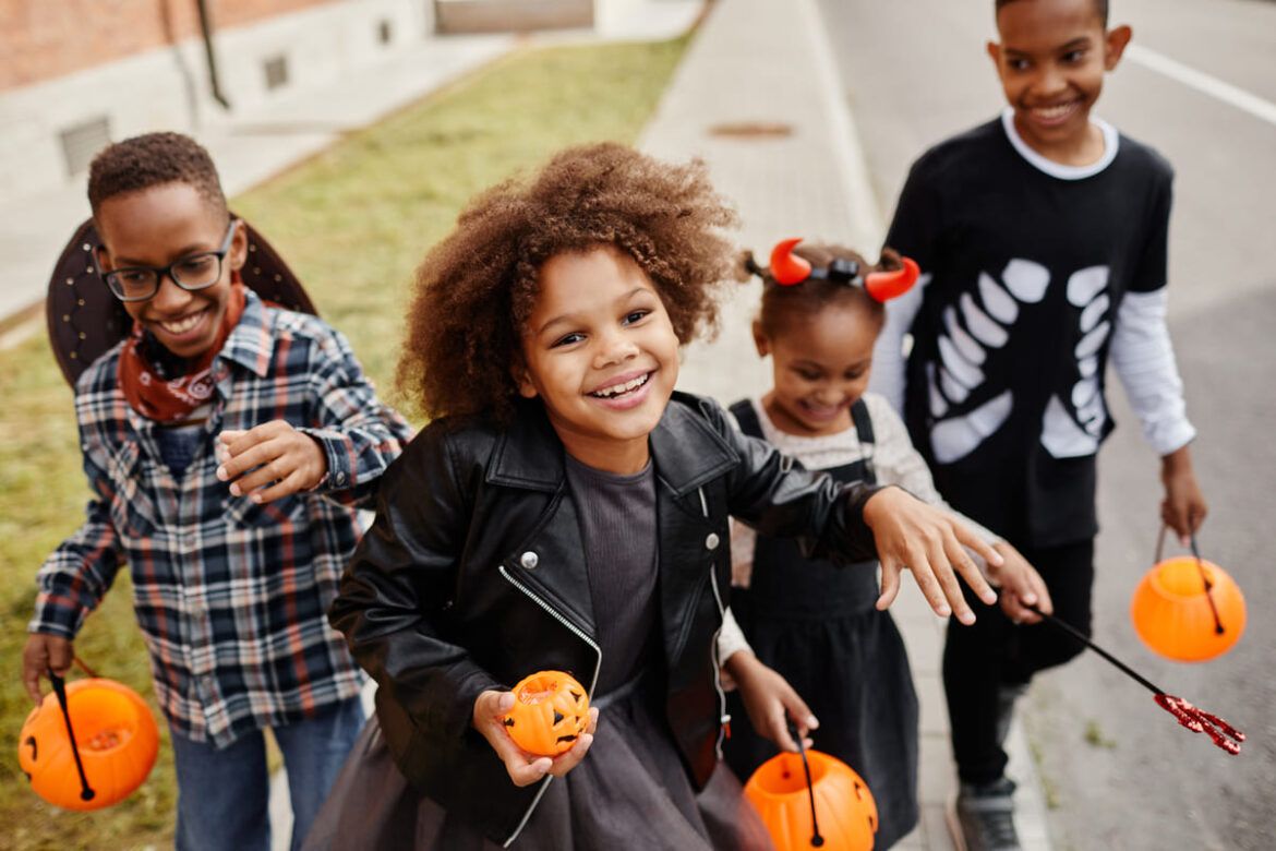 Crianças sorridentes vestidas com fantasias de Halloween e segurando abóboras de doces enquanto caminham pela rua.