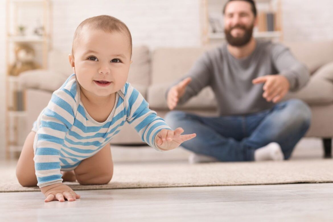 Bebê engatinhando no tapete e pai ao fundo.