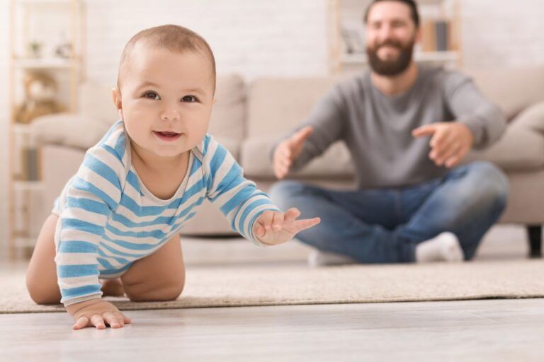 Bebê engatinhando no tapete e pai ao fundo.