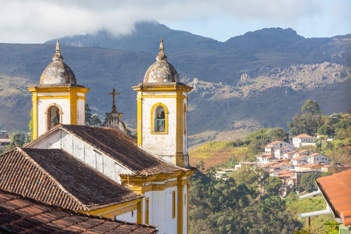 Vista de Ouro Preto com igreja à frente e a cidade abaixo. Há montanhas e muito verde.